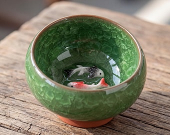 May include: A round ceramic tea bowl featuring a vibrant green crackle glaze finish. The interior of the bowl contains two small, three-dimensional fish figures, one coloured in black and white and the other in red and white, resting at the base.