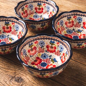 May include: A set of three ceramic bowls with a scalloped rim, featuring a cream-colored base decorated with a repeating pattern of red flowers, blue blossoms, and small yellow and black bees, finished with a dark blue trim along the edges.