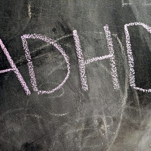 Puede incluir: Las letras ADHD están escritas con tiza de color rosa claro sobre una superficie de pizarra oscura y texturizada. Los trazos de tiza tienen un aspecto granulado y dibujado a mano, con marcas de tiza tenues y borrosas de fondo.