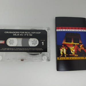 May include: A clear plastic cassette tape with a black label reading CRUSADERS FOR REAL HIP-HOP DÉJÀ VU - IT'S '82 and PROFILE, alongside a folded paper insert featuring a graphic of a yellow vehicle and people with the same title text.
