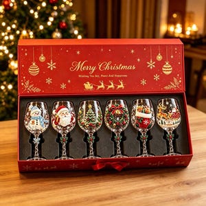 Op de afbeelding: Een set van zes wijnglazen van helder glas in een rode rechthoekige geschenkdoos met de tekst Merry Christmas Wishing You Joy, Peace And Happiness. Elk glas heeft een uniek kerstontwerp, waaronder een sneeuwpop, de kerstman, een boom, een krans, een kerstsok en een arrenslee.