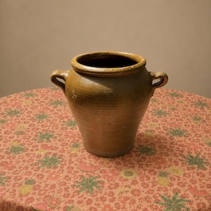 Op de afbeelding: Een bruine keramische pot met een brede ronde opening, een taps toelopend lichaam en twee kleine handvatten aan de zijkanten. De pot heeft een gladde, geglazuurde afwerking met subtiele kleurvariaties en een diepe, aardse tint.