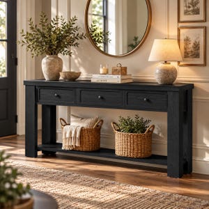 May include: A black wooden console table with three drawers and a lower shelf. The top surface holds a stone vase with greenery, a bowl, a candle, a small box, and two books titled The Kinfolk and At Home. Two woven baskets sit on the shelf.