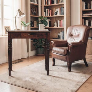 May include: A dark wood writing desk with four turned legs and a single drawer, paired with a brown leather armchair featuring a tufted backrest. The desk holds a metal lamp, a white ceramic mug, and a small potted plant on its surface.