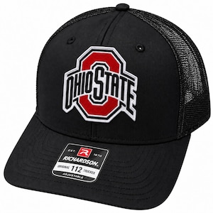 May include: A black trucker hat featuring a mesh back and a structured front panel. The front displays an embroidered Ohio State logo in red, white, and black. A rectangular sticker on the brim reads Richardson Original 112 Trucker Adjustable.