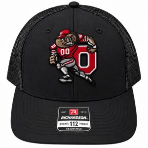 Könnte beinhalten: Eine schwarze Trucker-Cap mit Netzrücken und einem Vorderteil, das einen Cartoon-Charakter in einem roten Trikot mit der Nummer 00 vor einem großen roten Buchstaben O zeigt. Ein Aufkleber auf dem Schirm trägt die Aufschrift Richardson 112 Trucker.