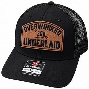 Op de afbeelding: Een zwarte truckerpet met een mesh achterkant en een bruine rechthoekige patch aan de voorzijde. De patch bevat de zwarte tekst OVERWORKED AND UNDERLAID. Een zwarte sticker op de klep toont de tekst EST. 1970 RICHARDSON ORIGINAL 112 TRUCKER ADJUSTABLE.