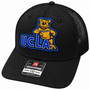 May include: A black trucker hat featuring an embroidered patch of the UCLA Bruins mascot in yellow and blue. The front panel displays the text UCLA in bold blue letters. A black sticker on the brim reads Richardson Original 112 Trucker Adjustable.
