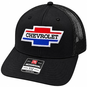 May include: A black trucker cap with a mesh back and a solid front panel featuring an embroidered Chevrolet bowtie logo in red, white, and blue. A rectangular sticker on the brim reads: EST. 1970 RICHARDSON ORIGINAL 112 TRUCKER ADJUSTABLE.