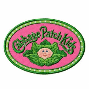 Könnte beinhalten: Ein ovaler, bestickter Aufnäher mit leuchtend rosa Hintergrund und grünem Rand, der mit weißen Punkten verziert ist. Der Schriftzug Cabbage Patch Kids ist in Grün über einem kleinen, pfirsichfarbenen Babygesicht in grünen Blättern gestickt.
