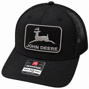 Puede incluir: Una gorra tipo camionero negra con un parche rectangular que muestra la silueta de un ciervo plateado saltando y el texto JOHN DEERE. El panel frontal es negro sólido y la parte trasera es de malla. Una pegatina en la visera dice RICHARDSON ORIGINAL 112 TRUCKER.