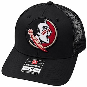 May include: A black trucker hat featuring a mesh back and a circular embroidered patch on the front. The patch displays a profile of a person with red face paint and a feather. A sticker on the brim reads Richardson Original 112 Trucker Adjustable.