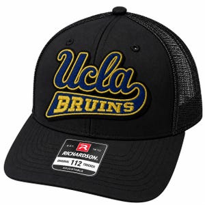 May include: A black trucker hat featuring a mesh back and an adjustable strap. The front panel displays an embroidered UCLA Bruins logo in blue and gold. A rectangular sticker on the brim reads Richardson Original 112 Trucker Adjustable.