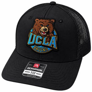 May include: A black trucker hat featuring an embroidered UCLA Bruins logo with a brown bear, blue text, and a yellow border. The front panel displays a rectangular sticker with the text EST. 1970 RICHARDSON ORIGINAL 112 TRUCKER ADJUSTABLE.