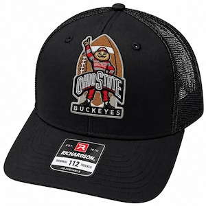 Puede incluir: Una gorra de camionero negra con parte trasera de malla y un parche bordado en el frente con el logotipo de la mascota de los Ohio State Buckeyes. El parche muestra una mascota en rojo y gris con el texto OHIO STATE BUCKEYES. Una pegatina negra en la visera dice RICHARDSON 112 TRUCKER.