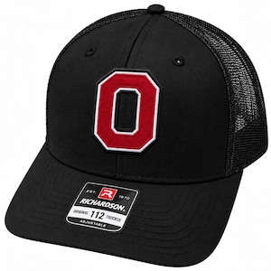 May include: A black trucker hat featuring a red and white embroidered letter O on the front panel. The hat has a mesh back, a curved brim, and a black sticker on the brim that reads Richardson Original 112 Trucker Adjustable Est 1970.