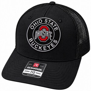 Puede incluir: Una gorra de estilo camionero negra con un parche circular en la parte frontal que muestra el texto OHIO STATE BUCKEYES y un logo O rojo. La gorra tiene una parte trasera de malla, una visera curva y una etiqueta negra RICHARDSON 112 TRUCKER.