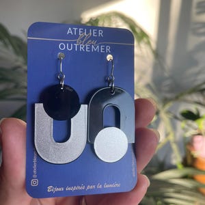 Könnte beinhalten: Ein Paar asymmetrische Ohrringe auf einer blauen Karte mit der Aufschrift ATELIER BLEU OUTREMER. Ein Ohrring zeigt einen schwarzen Kreis über einem silbernen U, der andere ein schwarzes U über einem silbernen Kreis. Beide hängen an silbernen Haken.