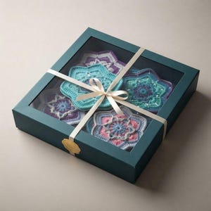 May include: A square dark teal gift box with a clear window lid containing four star-shaped crochet coasters in shades of purple, blue, pink, and teal. The box is tied with a cream-colored ribbon and features a small gold circular seal on the front.