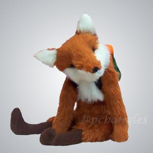 Könnte beinhalten: Ein Plüschfuchs mit orangefarbenem und weißem Fell, dunkelbraunen Pfoten und einem kleinen grünen Rucksack. Der Fuchs sitzt in einer Position mit geneigtem Kopf. Der Text @pcbangles ist auf der unteren rechten Seite des Spielzeugs zu sehen.