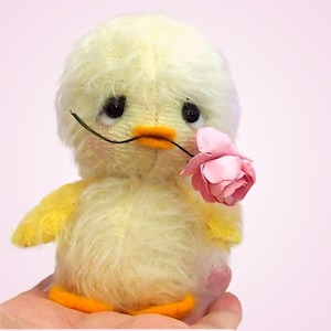 May include: A small, fluffy, pale yellow plush chick toy with dark, round eyes and an orange beak. The chick is holding a delicate, light pink artificial rose in its beak. The toy has a soft, textured appearance and rounded, stubby wings.