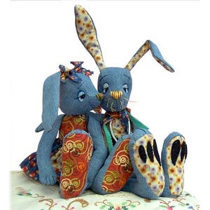 May include: Two blue fabric rabbit dolls with long ears. The rabbits feature orange fabric accents with gold swirl patterns on their feet and torsos. The ears have a floral pattern, and the faces have stitched eyes, noses, and whiskers.