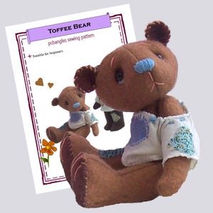 May include: A brown fabric teddy bear with a light blue nose, wearing a white top with blue floral and heart-shaped patches. The background features a sewing pattern document titled Toffee Bear by pcbangles with a small orange flower graphic.