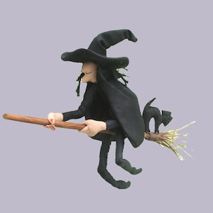 Op de afbeelding: Een stoffen heksenpop met een zwarte puntmuts en een lange zwarte mantel, rijdend op een houten bezemsteel. Een klein zwart kattenfiguurtje zit op de haren van de bezem. De pop heeft een lichte huid, donker haar en is in een vliegende houding.