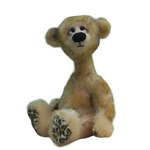 May include: A small, seated teddy bear with light tan, mottled fur and a long neck. The bear features a dark brown stitched nose, dark eyes looking upward, and distinct leopard print patterns on the soles of its feet and palms of its hands.