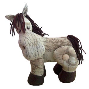 May include: A stuffed horse toy made of cream-colored fabric featuring a brown cursive script pattern. The horse has dark brown yarn for its mane and tail, and dark brown fabric covering its hooves. A small wooden button is visible on the hip.