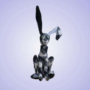 Può includere: Un coniglio di peluche realizzato in tessuto a quadri bianco e nero. Il coniglio presenta un orecchio lungo e dritto e un orecchio piegato, con piccoli occhi verdi e un naso cucito. Il corpo è composto da diversi pannelli di tessuto.