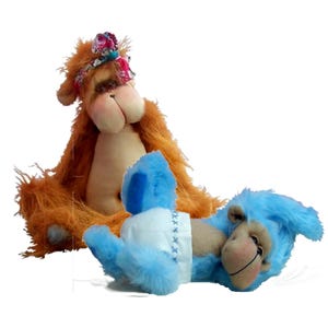 May include: Two plush monkey dolls. One is orange with long fur and a floral headband. The other is blue with long fur, lying down, and wearing a white nappy with blue cross-stitch detailing. Both have tan faces with stitched noses and mouths.
