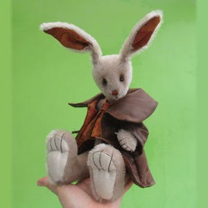 May include: A plush rabbit toy with long, upright ears featuring a brown paisley pattern on the inner side. The rabbit has light beige fur, dark eyes, and a brown nose, and is wearing a dark brown coat over an orange patterned vest.