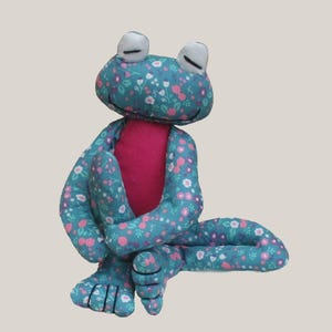 Könnte beinhalten: Ein Stofffrosch mit einem türkisfarbenen Stoffkörper, der mit einem Blumenmuster aus rosa und weißen Blüten bedruckt ist. Der Frosch hat einen einfarbigen, leuchtend pinken Bauch, weiße Augen mit schwarzen Sticklinien und lange, sitzende Gliedmaßen.