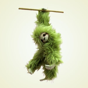 May include: A plush sloth toy covered in long, shaggy lime green faux fur. The sloth has a light-colored face with dark eyes and a stitched nose and mouth. It is hanging from a thin, horizontal wooden dowel by its front paws and claws.