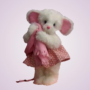 May include: A plush mouse toy with white fur, large pink-lined ears, and a pink nose. The mouse is wearing a red and white patterned dress and is holding a soft pink scarf with fringed ends. A thin, dark tail extends from the base of the figure.