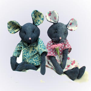 May include: Two stuffed mouse dolls made of dark blue denim fabric. The left mouse wears a teal shirt with a light floral pattern. The right mouse wears a white dress with a pink floral pattern and holds a single pink carnation flower.