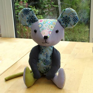 May include: A stuffed mouse toy featuring a light gray head with large ears patterned in blue and white floral designs. The face has black bead eyes and a stitched nose. The body is covered in a blue floral fabric with dark gray arms and a long green tail.
