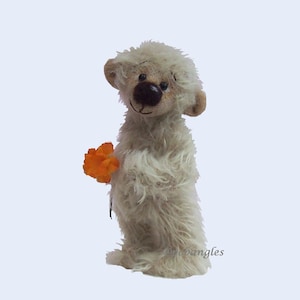 May include: A small, cream-colored plush bear with long, shaggy fur, a dark brown nose, and black eyes. The bear is standing upright and holding a small, bright orange flower in its paw. The text "@nebangles" is visible in the lower right corner.