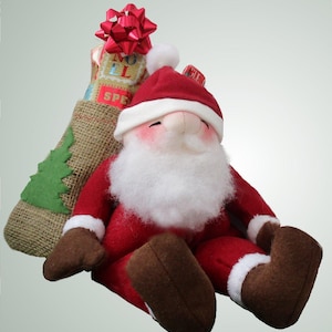 May include: A plush Santa Claus figurine wearing a red suit with white trim and brown boots, sitting beside a hessian sack decorated with a green felt tree and a red bow. The sack features text that reads NOEL and SPECIAL in gold lettering.