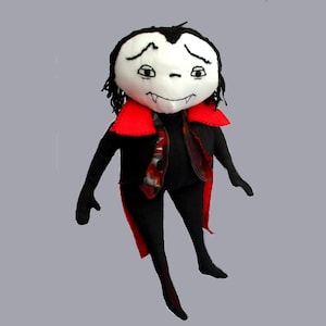 May include: A plush doll featuring a white face with embroidered black eyes, eyebrows, and fangs. The doll has black hair, a black suit, and a red-lined cape with a patterned inner fabric. The figure has long black arms and legs in a standing pose.