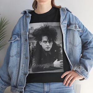 May include: A black t-shirt featuring a black and white photographic print of a person with voluminous dark hair wearing a leather jacket, layered under a light blue denim jacket with metal buttons and two chest pockets, paired with blue denim jeans.