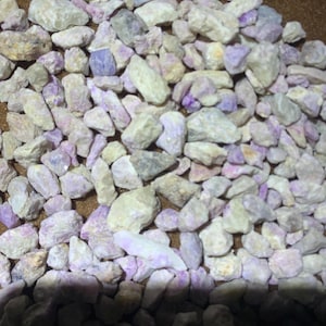 May include: A collection of small, raw, irregular mineral stones in shades of pale lavender, soft purple, and off-white. The stones have a rough, textured surface and vary in size and shape, creating a dense, scattered arrangement of light-colored, earthy, and pastel-toned rock fragments.