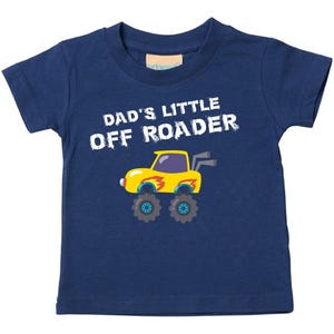 May include: A navy blue short-sleeved t-shirt featuring the white text DAD'S LITTLE OFF ROADER above a graphic of a yellow monster truck with flame accents, large black tires, and two grey exhaust pipes extending from the rear of the vehicle.