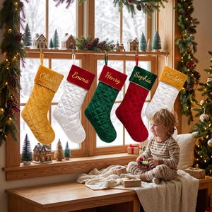 May include: Five quilted Christmas stockings hanging in a row, each featuring a personalised name embroidered in gold thread on the cuff. The stockings are coloured mustard yellow, white, dark green, red, and white with a mustard yellow cuff.