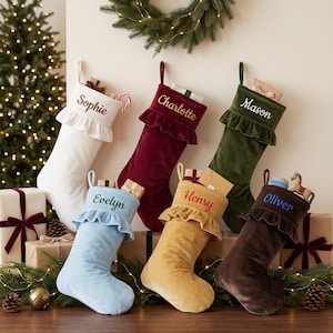 May include: Six velvet Christmas stockings with ruffled cuffs, each embroidered with a name: Sophie, Charlotte, Mason, Evelyn, Henry, and Oliver. The stockings come in white, burgundy, dark green, light blue, gold, and dark brown colours.