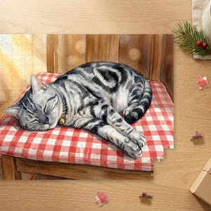 以下が含まれることがあります： 小さな金の鈴の首輪をつけた銀色のトラ猫が、赤と白のチェック柄のクッションの上で眠っているジグソーパズルのイラスト。木製の椅子の上に置かれ、周囲にはパズルのピースやギフトボックスが散らばっている。