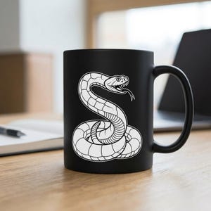 Op de afbeelding: Een matzwarte keramische mok met een witte lijntekening van een opgerolde slang met een geopende bek, waarbij giftanden en een gespleten tong zichtbaar zijn. De slang staat in het midden van de mok, die aan de rechterkant een gebogen handvat heeft.