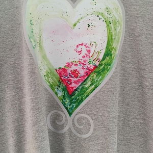Könnte beinhalten: Ein graues meliertes T-Shirt mit einer großen, mittigen Herzgrafik. Das Herzdesign zeigt aquarellartige Farbspritzer in Hellgrün, Weiß und leuchtendem Pink sowie ein dekoratives weißes Wirbelmuster am unteren Ende des Motivs.