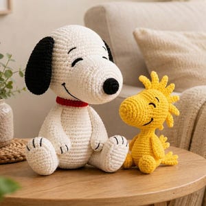 May include: A white and black crocheted Snoopy figure with a red collar sitting next to a smaller yellow crocheted Woodstock figure. Both figures have stitched facial features and are crafted with a visible yarn texture in a seated position.